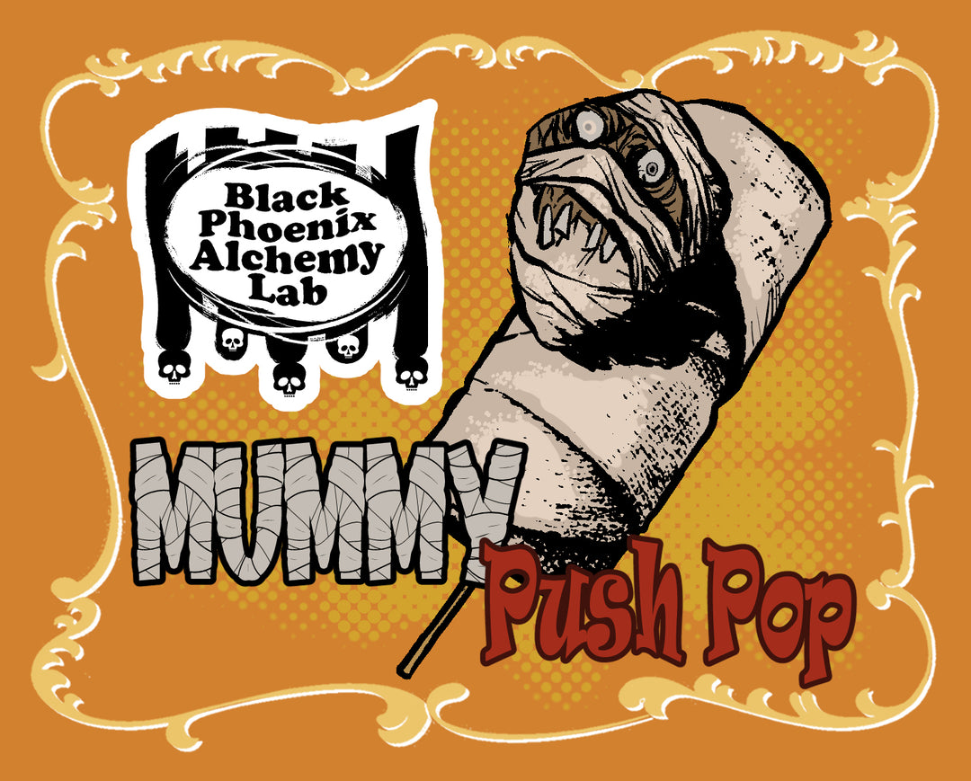 Black Phoenix Alchemy Lab Mummy Push Pop illustration: a bandage-wrapped push pop treat with a snarling, wide-eyed mummy face bursting through the linen coating, on an orange halftone background with an ornate gold border and the BPAL skull-topped logo.
